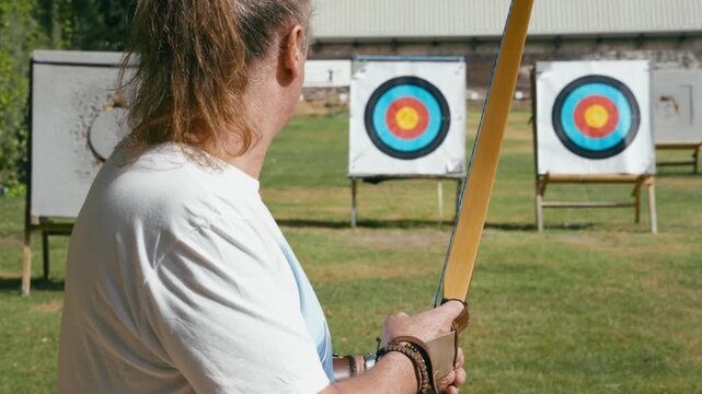 Senior man with long hair training his archery skills. Archer holding a longbow, aiming and releasing an arrow towards a distant target