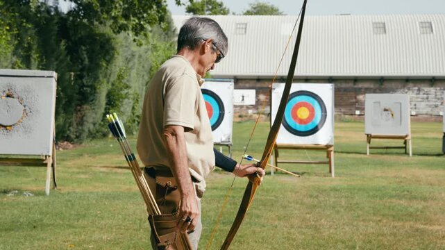 Older man training with a longbow, aiming and shooting an arrow at a target. Archer practicing on a green field during a sunny day