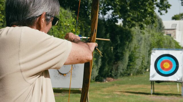 Mature man with gray hair concentrating and aiming with a wooden bow. Archer focusing on the bullseye during an outdoor archery practice