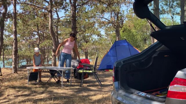 A mother and her young son set up a table at their campsite next to an open car trunk in a pine forest. This peaceful outdoor family adventure feels full of joy and active togetherness.