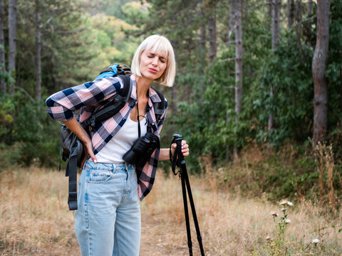 Woman hiker experiencing back pain while hiking in  forest, resting with hiking pole and looking worried after physical effort.