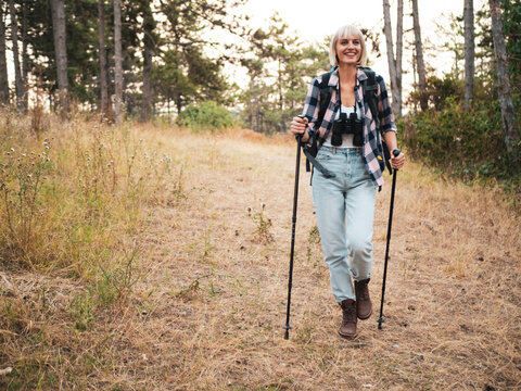 Carefree woman hiker walking on mountain footpath during hiking, holding hiking pole and carrying backpack, enjoying nature, adventure, travel and relaxation.