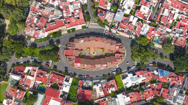 Top-down aerial timelapse of cars circulating around an oval residential roundabout surrounded by red tile rooftops and green trees