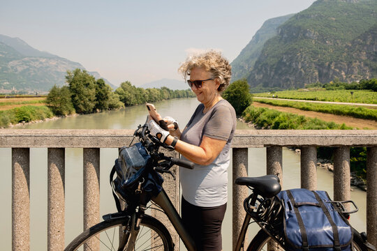 Senior woman cyclist checking phone by Adige River in Italy