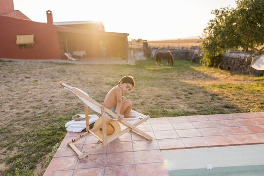 Boy climbing on deck chair by pool at country house in summer