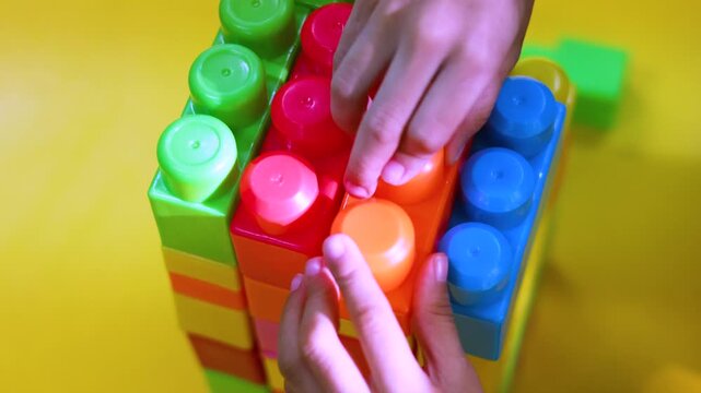 Close up 4k video of a toddler's hands artfully assembling colorful blocks. It highlights the benefits of constructive play in developing fine motor skills and encouraging early engineering sparks.