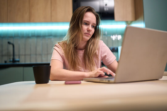 Freelance woman typing on laptop at home kitchen table with cup