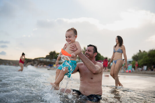 Father holding toddler son splashing water at beach