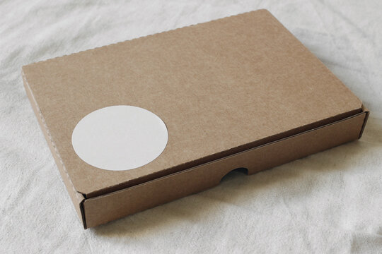 Cardboard gift box mockup with closed lid. Blank white circular sticker template for logo, text placement. E-commerce packaging shipping concept. Blurred beige textile background, top view
