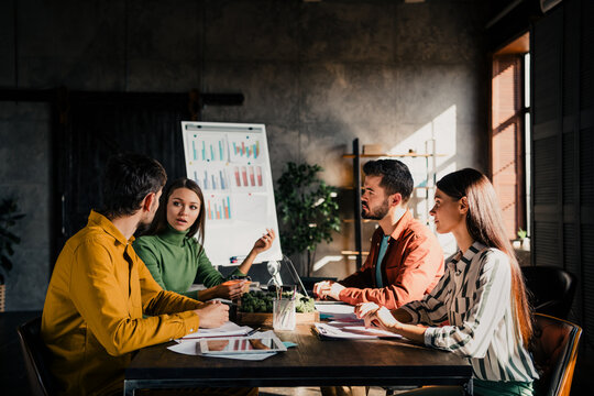 Business team meeting around a table discussing charts and strategy in a modern office with laptop, flip chart and natural light