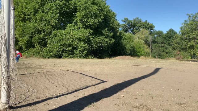 A soccer goal casts a long shadow across a dry field while a person walks near green trees. The sunny outdoor park feels peaceful and nostalgic.