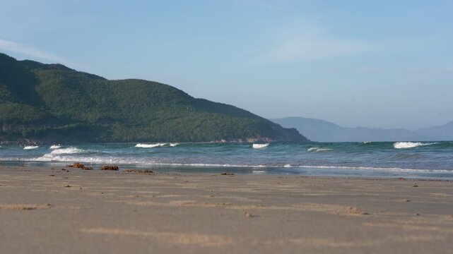 Serene Coastal Landscape: Gentle Waves Lapping on Sunlit Sands with Lush Hills in the Background Under a Clear Blue Sky