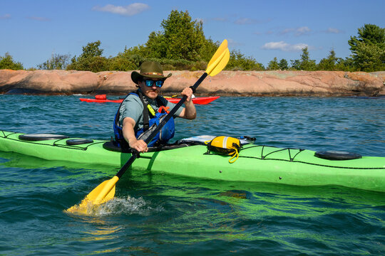 Sea kayaker in a wide-brim hat paddles a green kayak with a yellow paddle past pink granite on Georgian Bay, Ontario, Canada