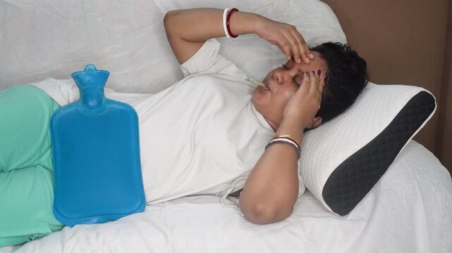Indian young sick woman checking fever with thermometer during menstrual periods. Using hot water bag to relive cramp or pain, while lying on sofa at home. Fever and illness concept.