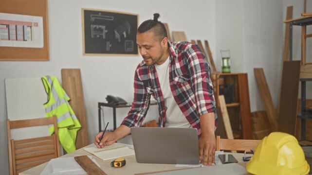 Man carpenter writing measurements and notes by laptop at a woodworking table with tape measure and hard hat in a studio; concentration planning.