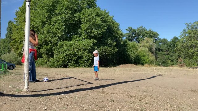 A loving mother gently kicks a soccer ball toward her happy young son. The joyful child watches excitedly on a sunny dirt field under a bright blue sky.