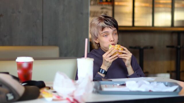 A Caucasian teenage girl sits in a welcoming cafe, enjoying a juicy burger as she savors every flavorful bite in the sunlit, cheerful atmosphere around her. Slow motion 4k video.