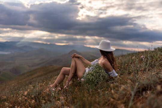 Young woman wearing white hat rests on grassy hillside enjoying scenic mountain landscape view at dusk