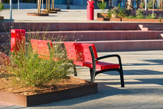 Red public bench sits beside planted beds on modern urban plaza. Modern outdoor seating and landscape design for public space in city