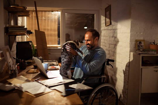 Man in wheelchair lifting cat at home office desk