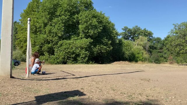 A young boy squats beside a woman next to a soccer ball by the goal post. They share a tender, loving moment on a sunny dirt field surrounded by green trees.