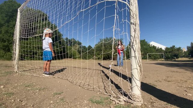 A focused little boy waits at the goal while a happy woman prepares to kick the ball. They enjoy an energetic game on a sunny outdoor field filled with carefree joy.