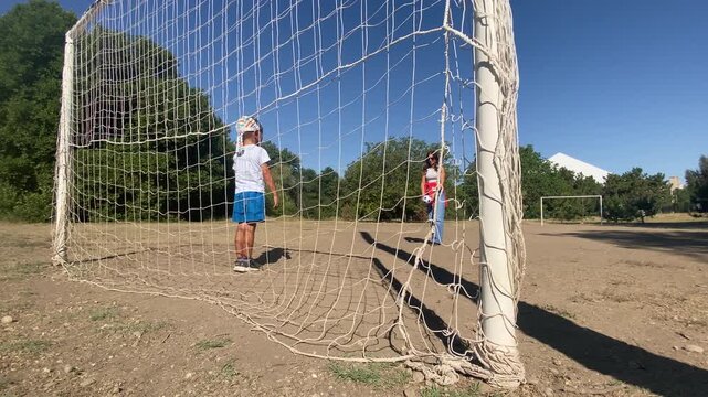 A young boy waits inside a soccer goal while a woman holds a ball to play. They enjoy a fun football game together on a sunny outdoor dirt field. The atmosphere feels happy and warm.