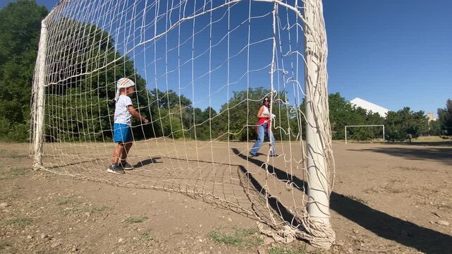 A small boy inside a soccer goal points across the net to a woman holding a football. They play a cheerful game on a sunny dirt field. The atmosphere feels happy and warm.
