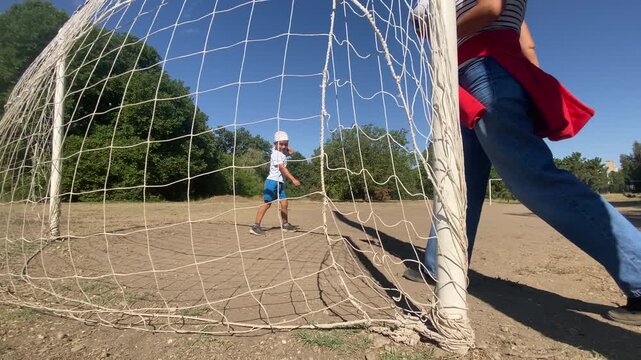 A woman holds a soccer ball and walks through a goal net while a child plays nearby. They share a playful football game on a sunny dirt pitch. The outdoor scene feels warm and joyful.