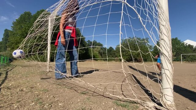 A woman bends down inside a soccer goal while a little boy walks nearby. They play a football game on a sunny dirt pitch. The scene feels warm, happy, and playful.