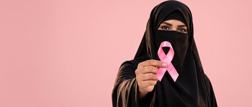 Breast cancer awareness concept. Arab Muslim woman in a black niqab holding a pink ribbon on a pink background, long banner with copy space.