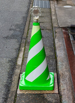 Close-up of a green and white striped traffic cone equipped with a transparent warning light, positioned beside a curb on a narrow paved urban street
