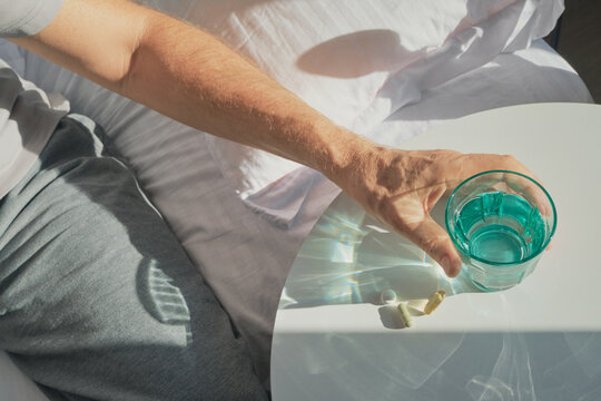 Man reaching for medication and water on bedside table, illuminated by morning sunlight