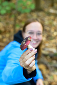 A close-up view of a hand holding a wild mushroom with a red cap in an autumn forest in Paris, France. A smiling woman is softly blurred in the background.