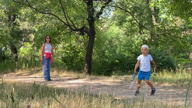 A loving mother and her joyful young son play badminton. The cheerful boy walks along a dirt path in a leafy green park. They share a sweet and peaceful moment.
