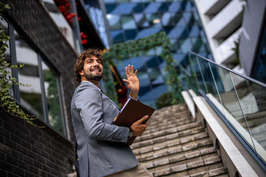 Smiling businessman holding clipboard and waving hand on outdoor stairs