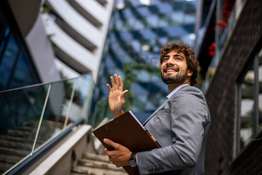 Low angle portrait of businessman waving hand with clipboard outside office
