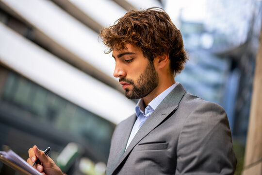 Young man in suit writing notes on clipboard outdoors in urban environment