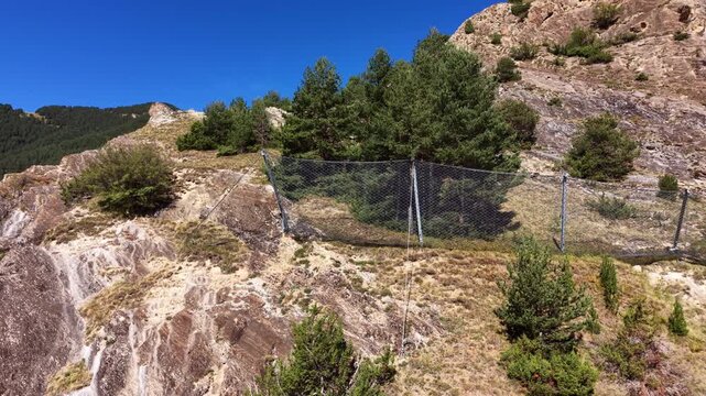Metal mesh rockfall barrier net installed on a steep rocky mountain slope with scattered pine trees under a clear blue sky, erosion control infrastructure in alpine terrain