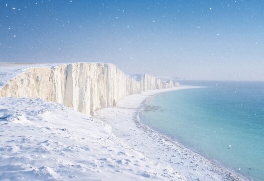 Dramatic winter snowfall covers white chalk cliffs above a stormy sea, with crashing waves, rocky shoreline, dark clouds, and rugged coastal scenery.