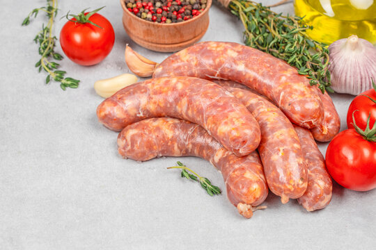 Raw sausages arranged with tomatoes, garlic, thyme, peppercorns, and oil on a light stone surface, fresh ingredients for cooking, culinary food still life