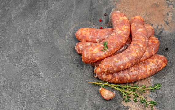Raw seasoned sausage links arranged with garlic, herbs, red peppercorns, and black pepper on a dark textured slate surface, overhead food still life