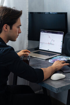 Young man in black shirt analyzes financial data on laptop screen while sitting at desk with cash and a cup of coffee in a modern home office setting