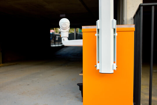 Open orange parking barrier control box with visible motor gears and wiring beside lowered striped barrier arm on urban roadside in daylight near brick wall and paved curb