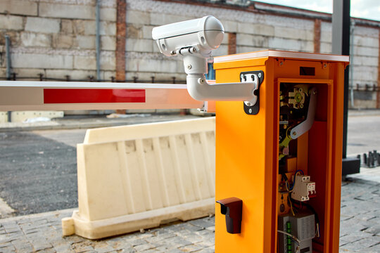 Open orange parking barrier control box with visible motor gears and wiring beside lowered striped barrier arm on urban roadside in daylight near brick wall and paved curb