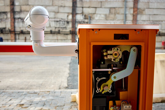 Open orange parking barrier control box with visible motor gears and wiring beside lowered striped barrier arm on urban roadside in daylight near brick wall and paved curb