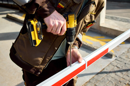 Closeup of construction worker using cordless drill to fasten red and white metal barrier on street-side sidewalk during daylight with visible hand driving screws into guardrail