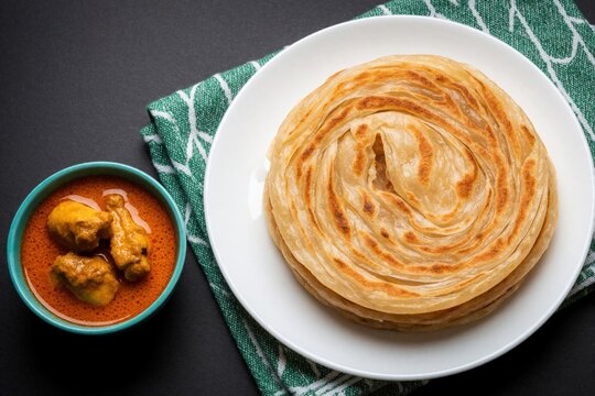 Tasty Indian parotta bread served with spicy chicken curry in small bowl. Flaky layered flatbread sits on white plate next to meat gravy. Traditional South Indian cuisine meal on dark table with