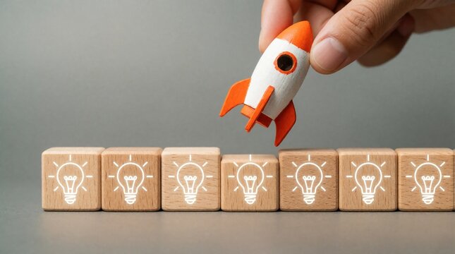Hand places small rocket toy above wooden blocks with light bulb icons. Business concept shows startup launch, product release, new idea deployment. Successful project growth starts with innovation,