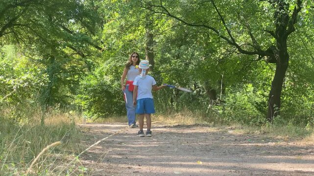 A mother and son stand ready to play badminton. They hold rackets along a dirt path in a lush green forest. The sunny scene creates a warm and happy feeling.